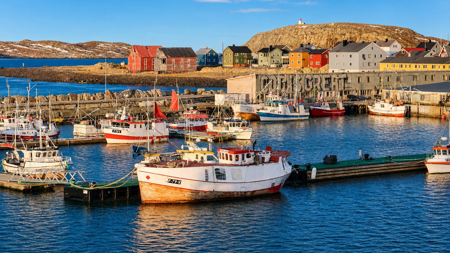 Colourful fishing boats and painted harbour houses below the lighthouse hill at Vardo, Norway.