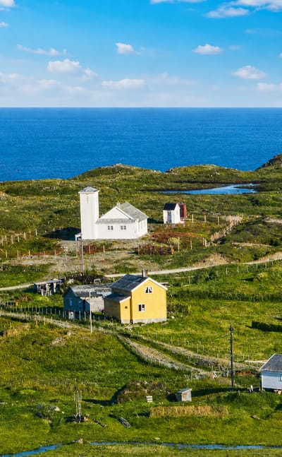 Coastal hamlet of Vuonnabahta, Norway, with white and yellow houses on green tundra above a deep-blue sea.