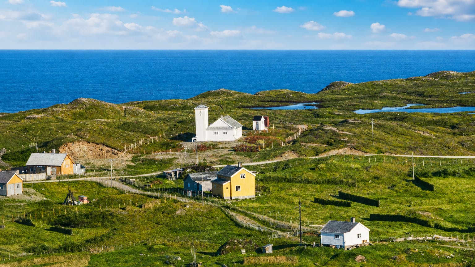 Coastal hamlet of Vuonnabahta, Norway, with white and yellow houses on green tundra above a deep-blue sea.