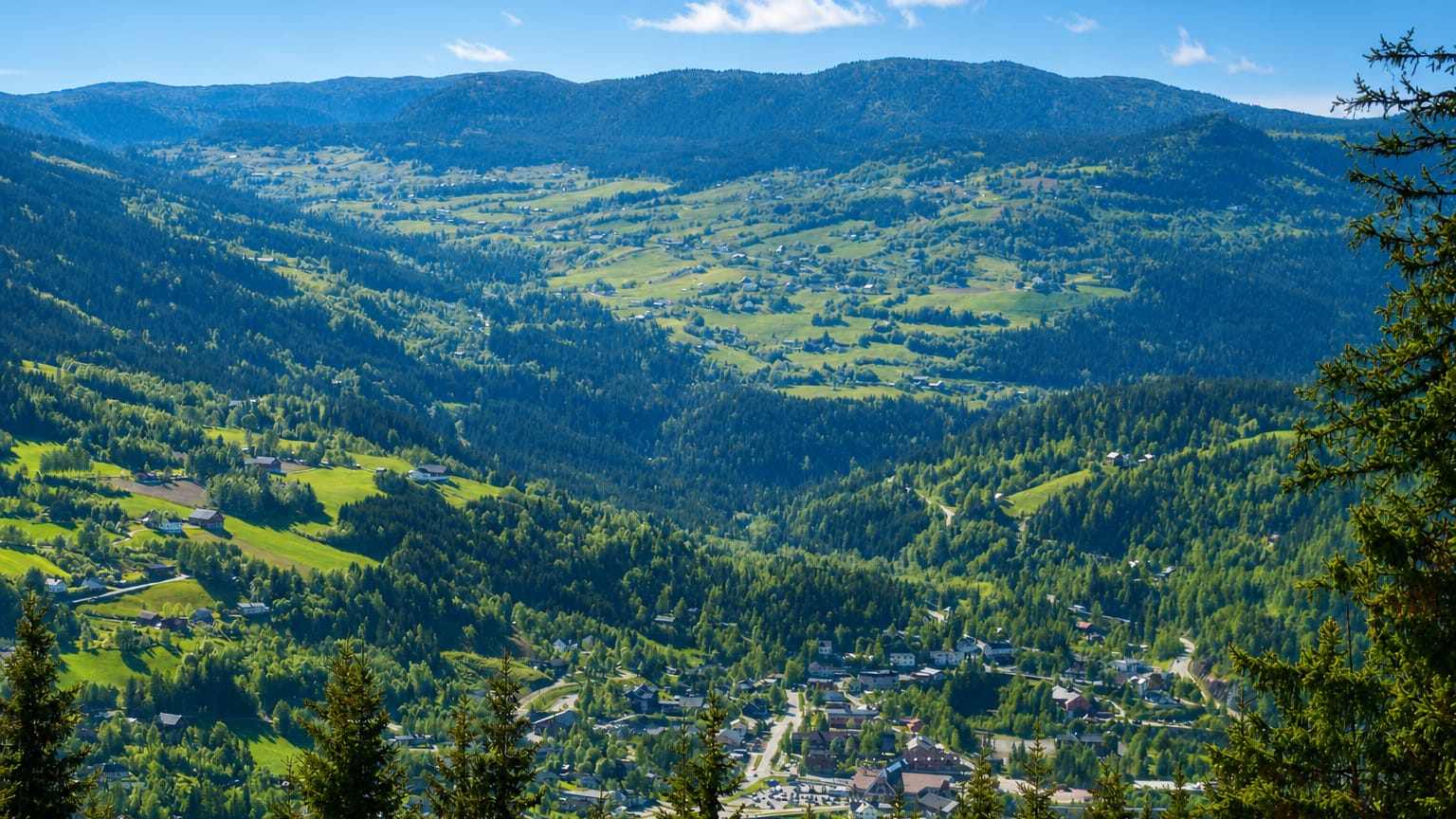 Bagn village nestled on the floor of a green, forested Valdres valley ringed by wooded hills under a blue summer sky.