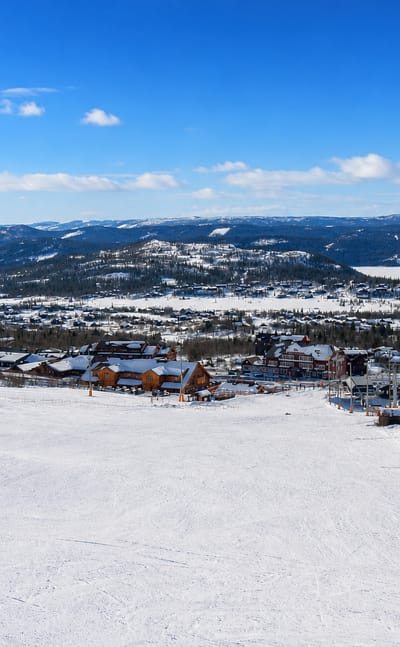 Snowy ski slopes, a lift and wooden lodges of Beitostølen, Norway, below forested hills and distant snow-capped mountains