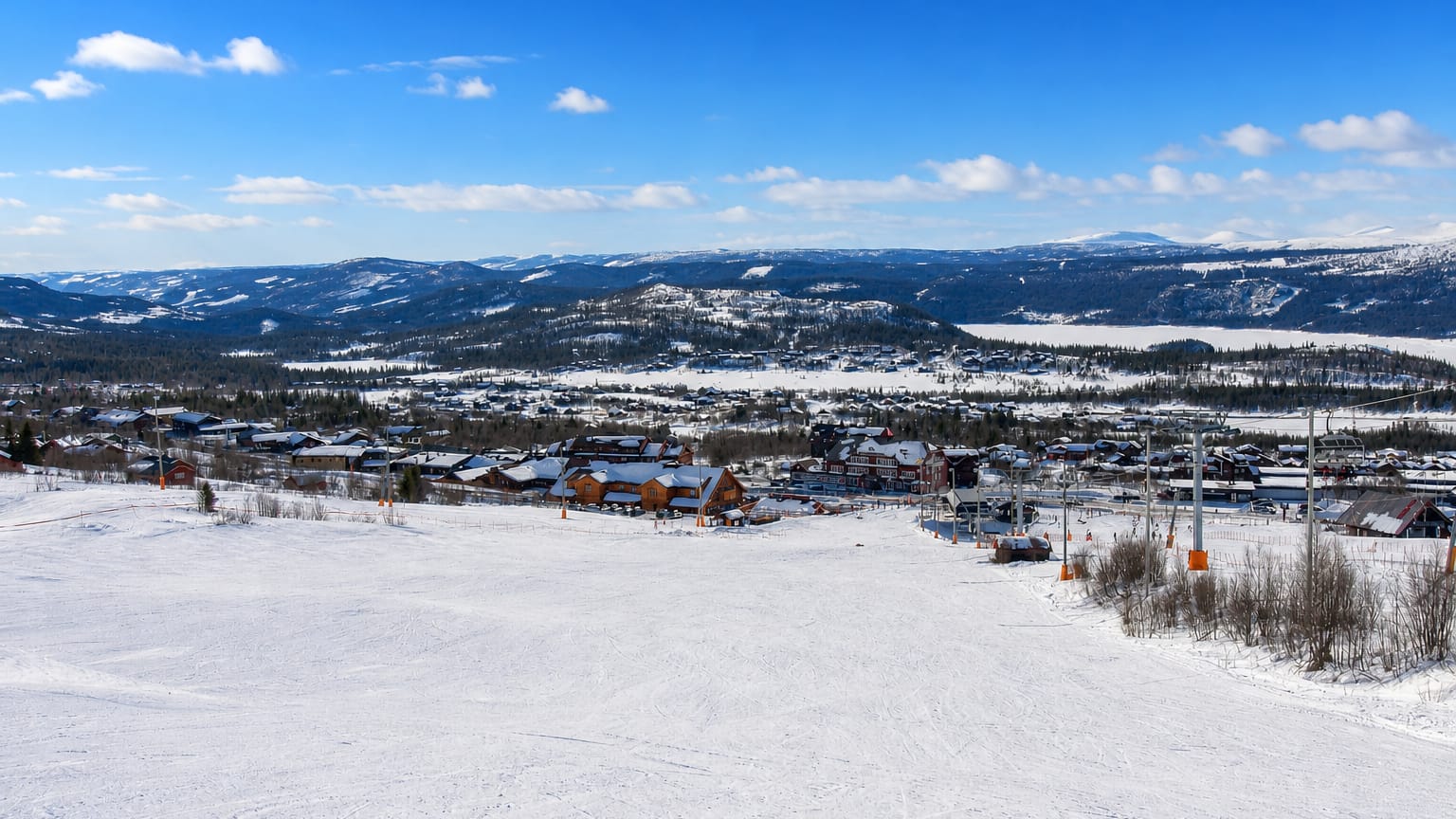 Snowy ski slopes, a lift and wooden lodges of Beitostølen, Norway, below forested hills and distant snow-capped mountains