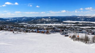 Snowy ski slopes, a lift and wooden lodges of Beitostølen, Norway, below forested hills and distant snow-capped mountains