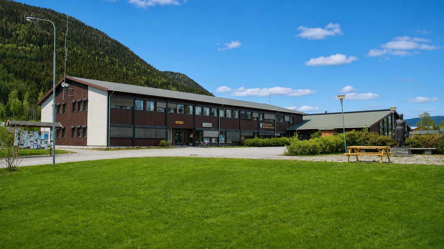 Brown-timber municipal building at Bergset with a lawn, statue and forested mountain rising behind