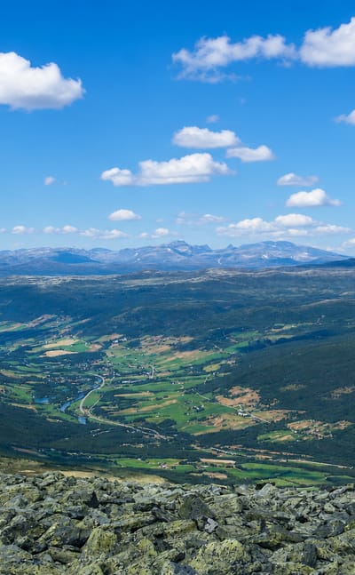Dovre's broad green valley with a winding river between forested mountains under scattered clouds, seen from a rocky summit