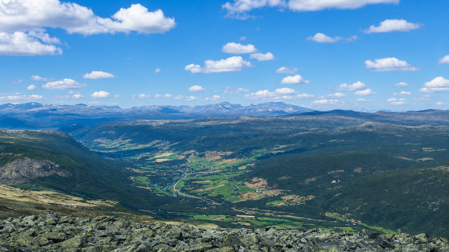 Dovre's broad green valley with a winding river between forested mountains under scattered clouds, seen from a rocky summit