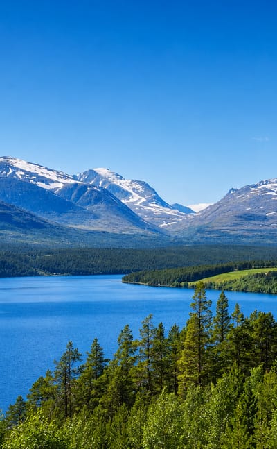 Snow-capped Rondane peaks rising above a forested blue lake near Folldal, Norway.