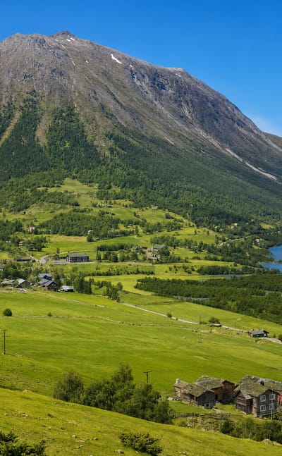Green mountain valley at Fossbergom, Norway, with scattered farms and turf-roofed buildings beside a lake below a steep peak.