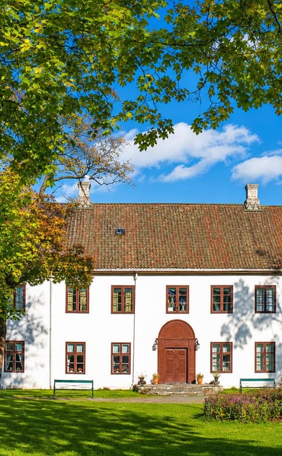 Gjøvik's white historic manor house with red-trimmed windows and a tiled roof amid autumn trees and lawns.