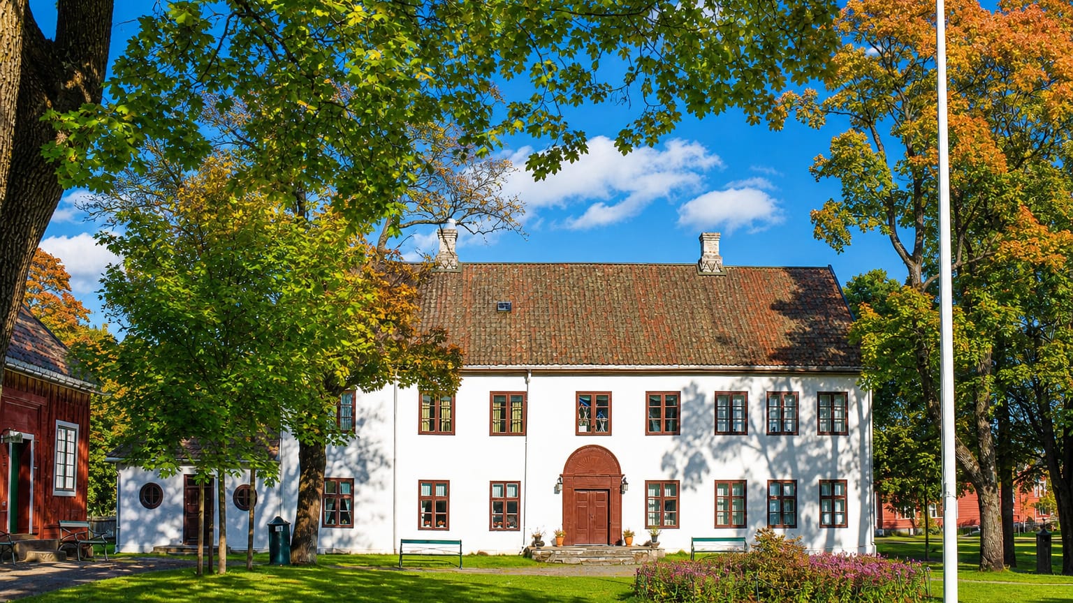 Gjøvik's white historic manor house with red-trimmed windows and a tiled roof amid autumn trees and lawns.