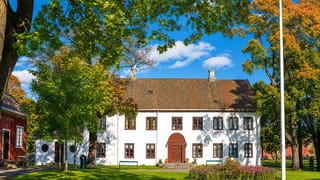 Gjøvik's white historic manor house with red-trimmed windows and a tiled roof amid autumn trees and lawns.