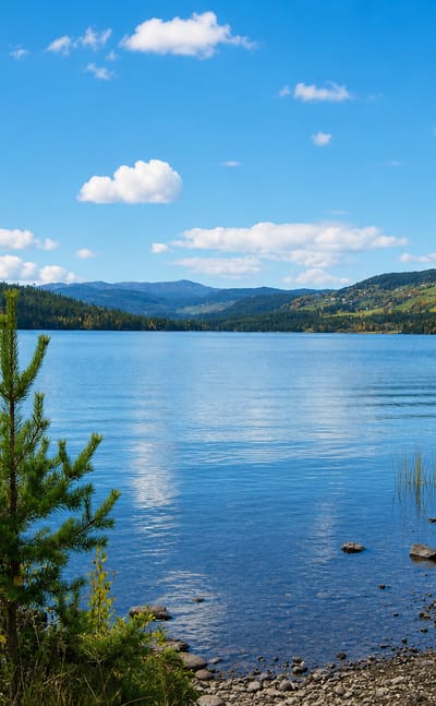 Calm blue lake ringed by forested hills and distant mountains at Heggenes, Norway, with a small boat and autumn shoreline.