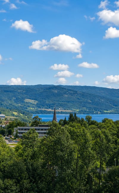 Hov village beside Randsfjorden lake, a church spire rising among low buildings amid forested hills under a blue summer sky