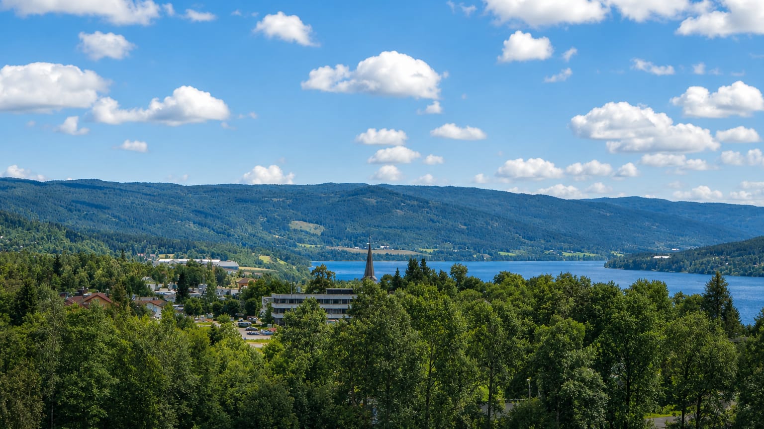 Hov village beside Randsfjorden lake, a church spire rising among low buildings amid forested hills under a blue summer sky