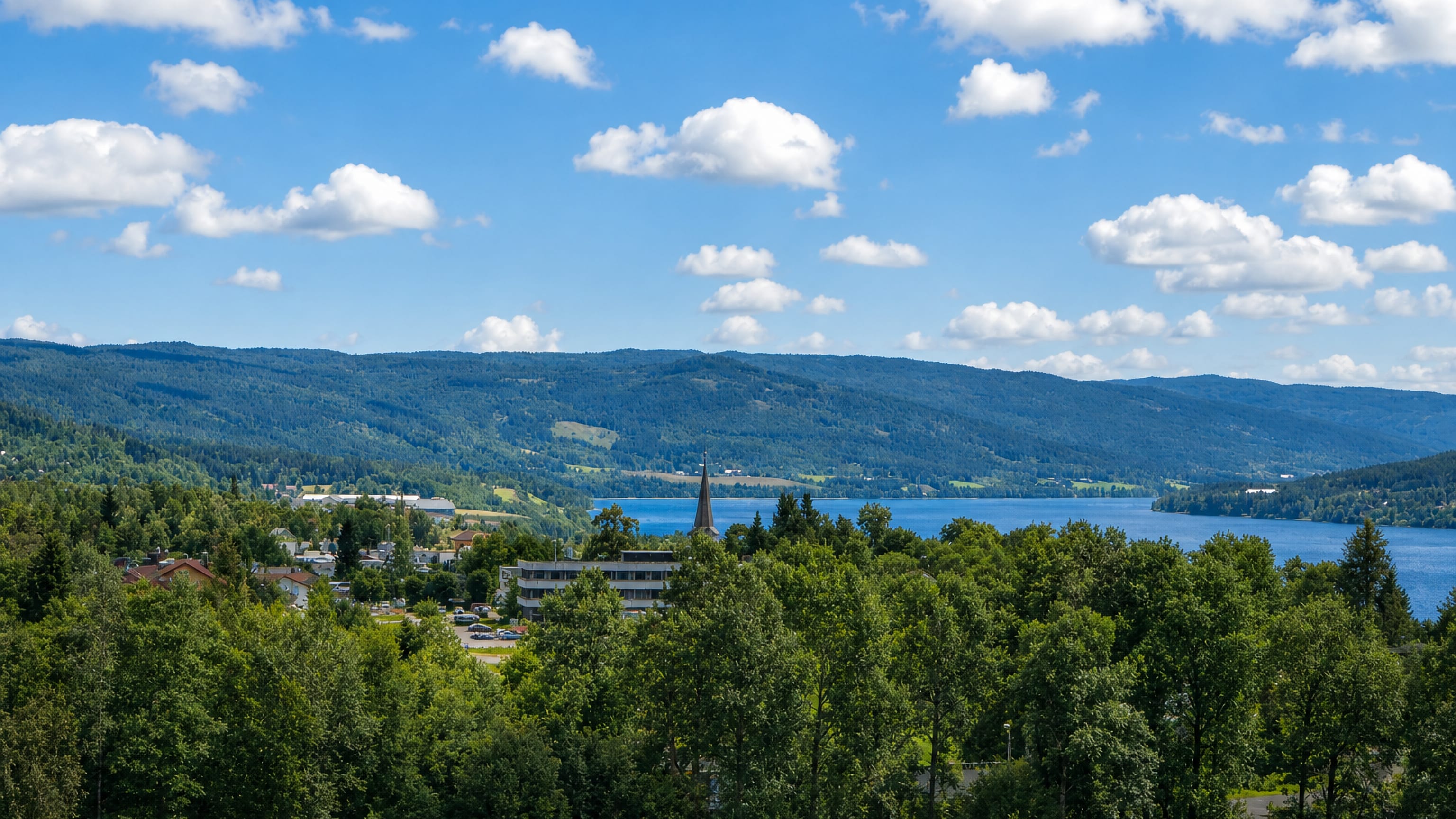 Hov village beside Randsfjorden lake, a church spire rising among low buildings amid forested hills under a blue summer sky