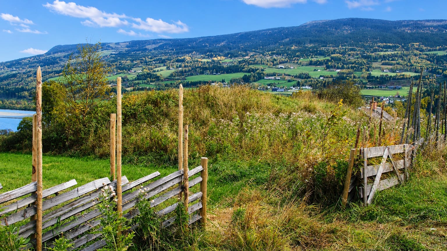 View over Hundorp's Gudbrandsdalen valley with farmland, a river and forested hills, framed by a traditional wooden rail fence