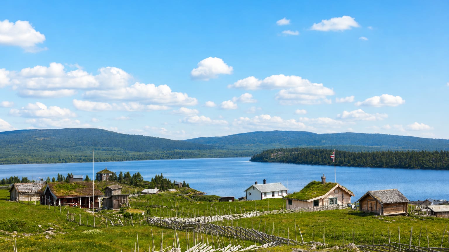 Traditional turf-roofed cabins and a Norwegian flag on grassy slopes above Sjusjøen lake, with forested hills beyond.