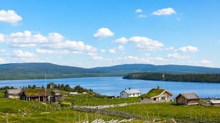 Traditional turf-roofed cabins and a Norwegian flag on grassy slopes above Sjusjøen lake, with forested hills beyond.