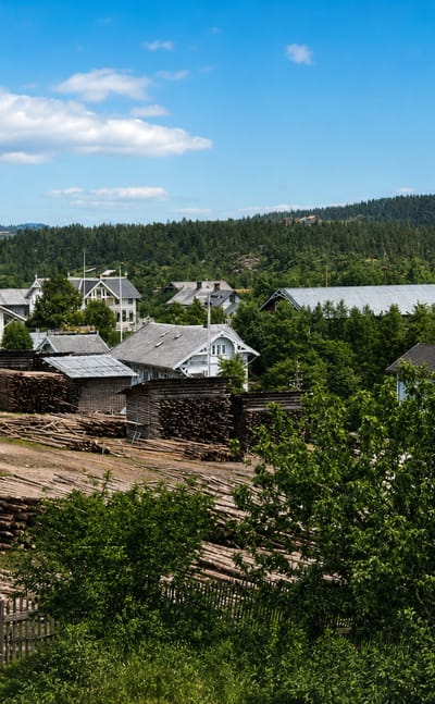 Skotterud village in Eidskog, Norway, with white wooden houses, stacked timber logs and forested hills above a distant lake.