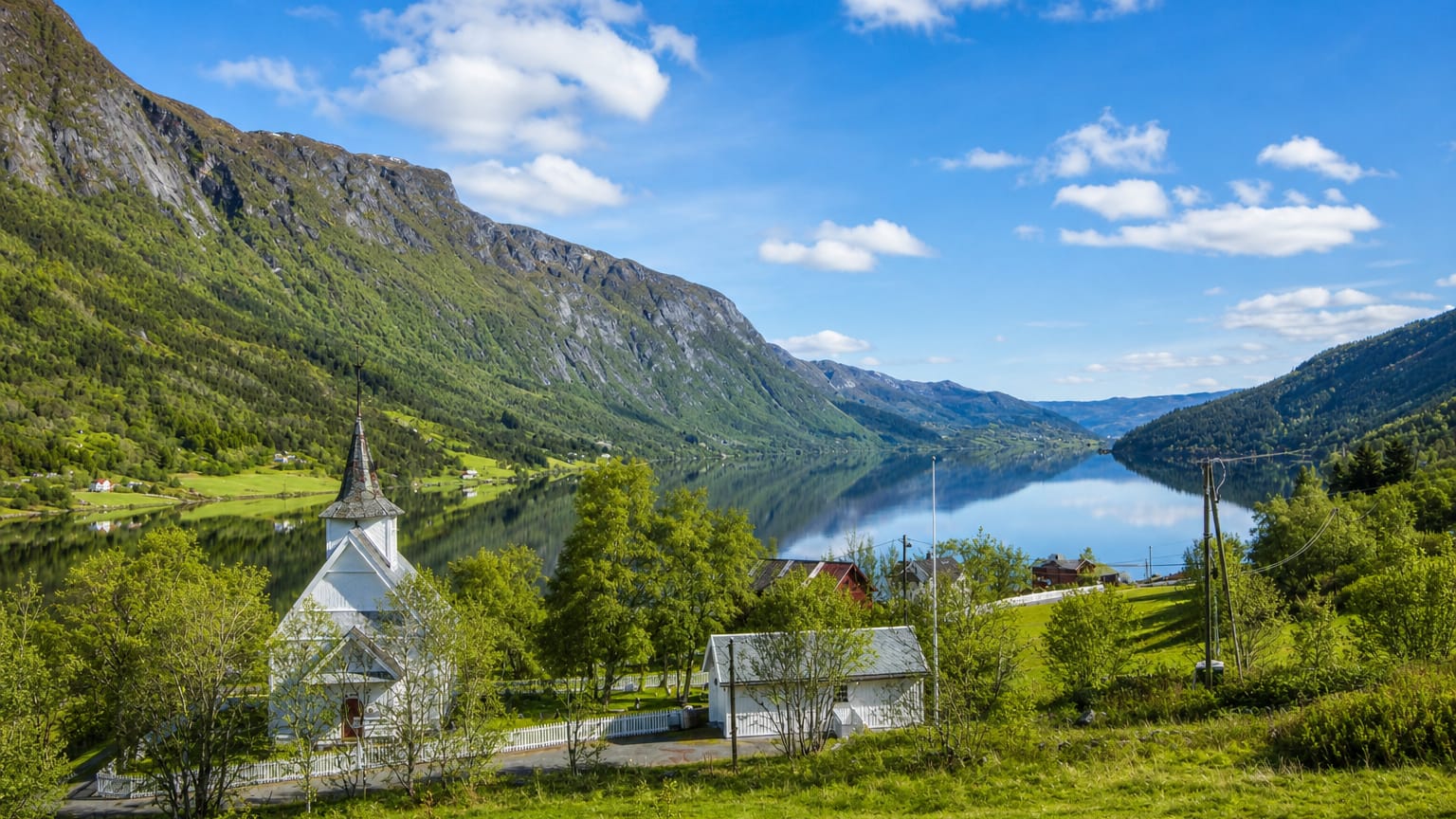White wooden church beside a calm lake ringed by forested mountains in Vang, Norway.