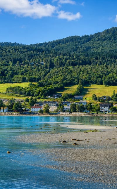 Fjord-side village of Eidsvåg, Norway, scattered houses and green meadows below forested hills by calm turquoise water