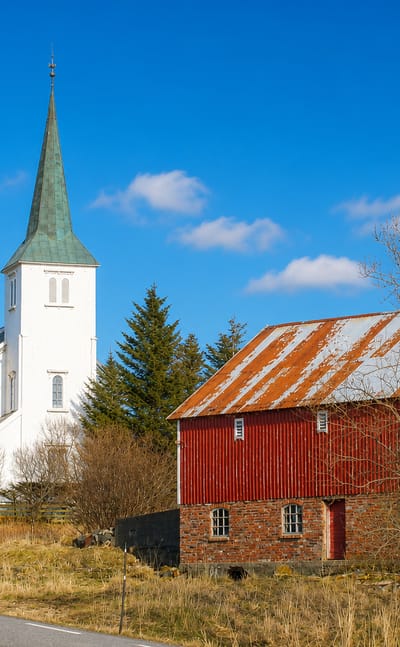 White church with a green spire beside a red timber barn under a blue sky in Hopen, Norway.