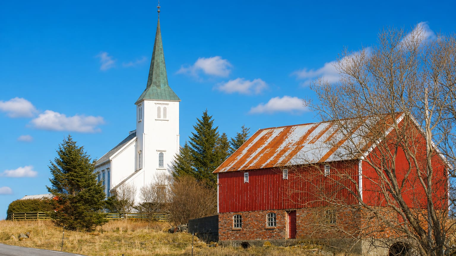 White church with a green spire beside a red timber barn under a blue sky in Hopen, Norway.