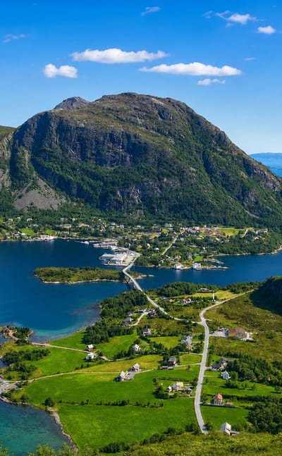 Aerial view of Midsund's island village beneath a green mountain, ringed by the blue Romsdalsfjord under a bright sky.