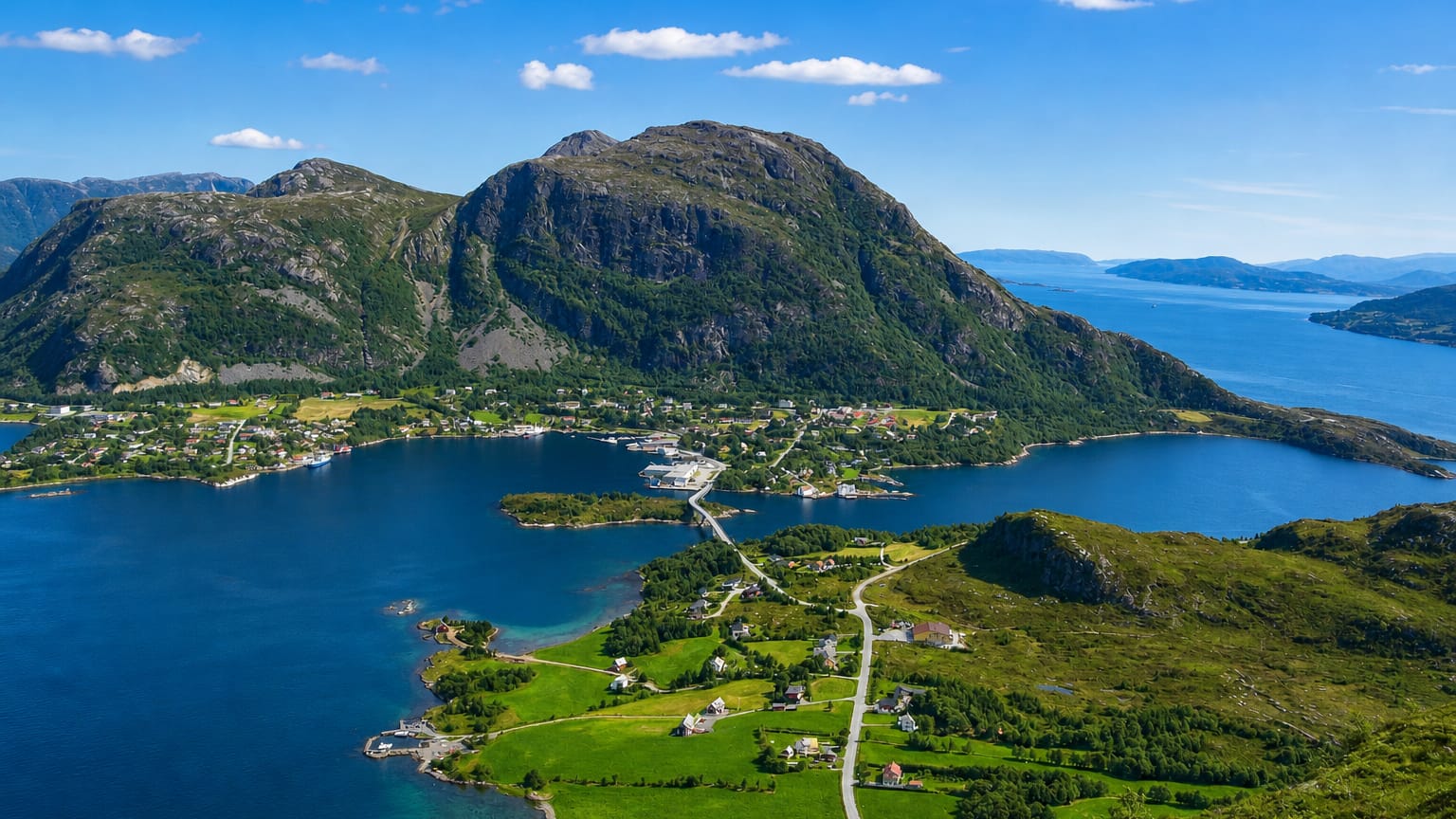 Aerial view of Midsund's island village beneath a green mountain, ringed by the blue Romsdalsfjord under a bright sky.