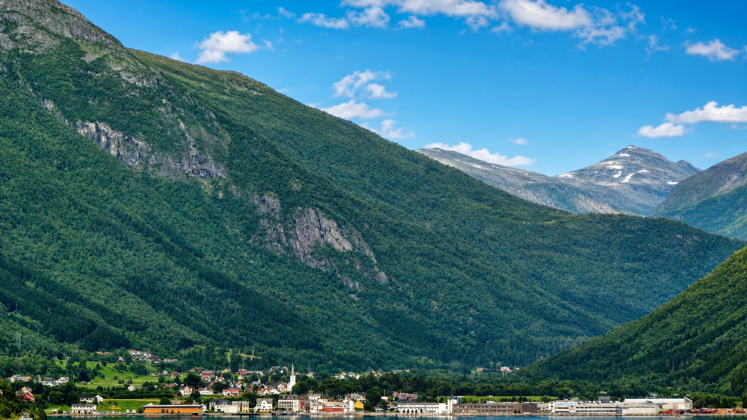 Stordal settlement along a blue fjord shore below steep forested mountains and distant snow-capped peaks, Norway.