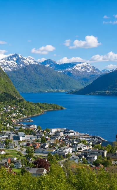 Volda, Norway, spread along a fjord shore below snow-capped Sunnmore peaks with a church tower and ferry quay.