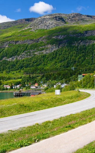 Winding road along the fjord at Bogen, Nordland, with red houses below a green forested mountain under a blue sky.