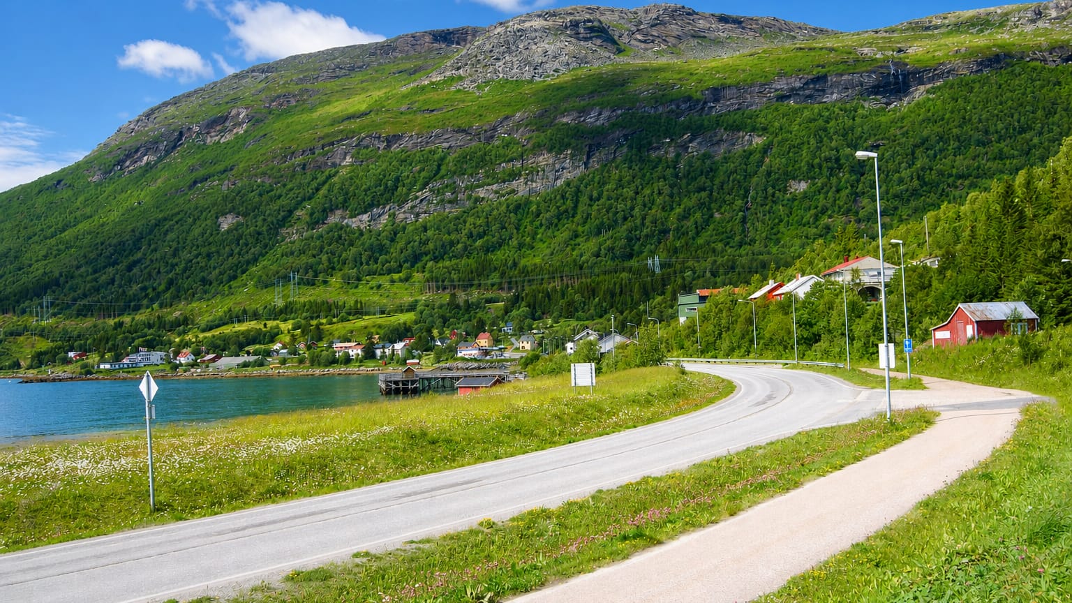Winding road along the fjord at Bogen, Nordland, with red houses below a green forested mountain under a blue sky.
