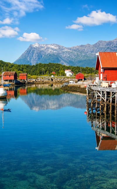 Red wooden boathouses on stilts and moored white boats in the calm, clear harbor at Inndyr, Norway, below coastal mountains.