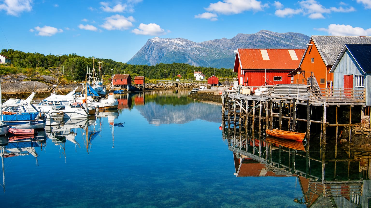 Red wooden boathouses on stilts and moored white boats in the calm, clear harbor at Inndyr, Norway, below coastal mountains.