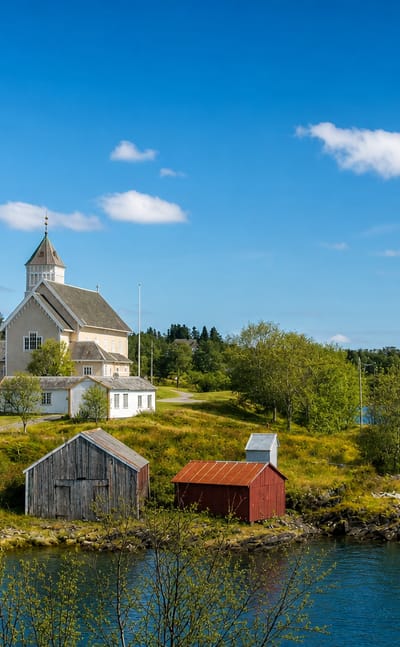 White wooden church on a green headland above the calm fjord at Leinesfjorden, Nordland, with red boathouses by the shore.