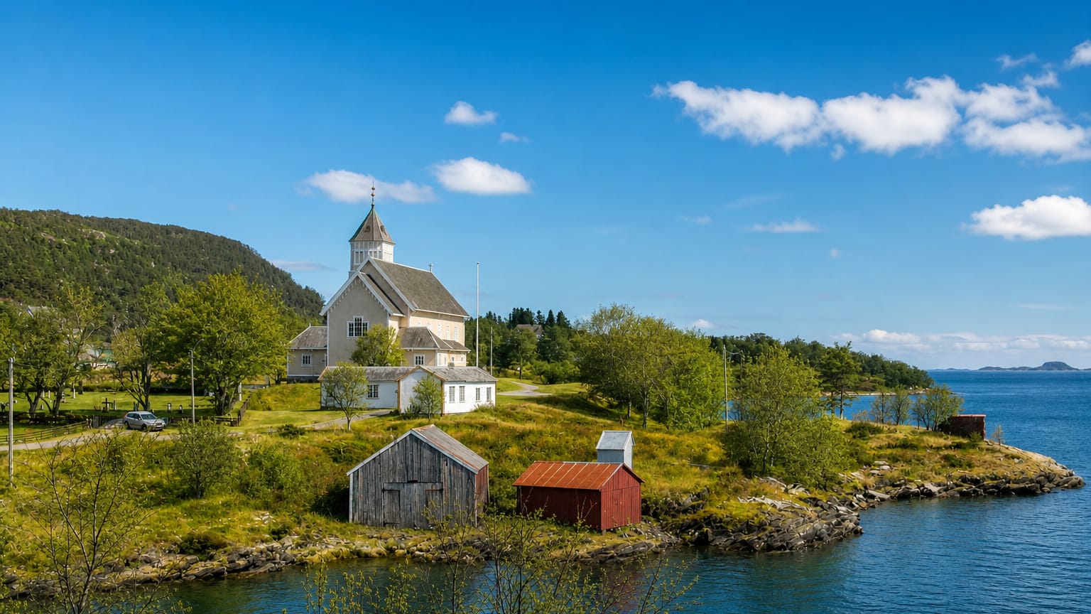 White wooden church on a green headland above the calm fjord at Leinesfjorden, Nordland, with red boathouses by the shore.
