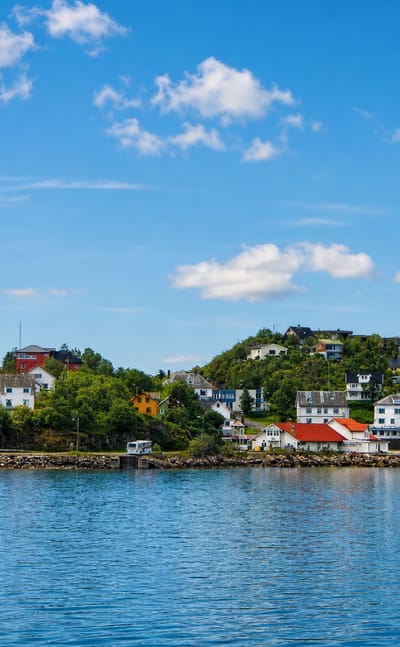 Waterfront of Lødingen, Norway, with white red-roofed houses along the shoreline below a forested hill under blue sky