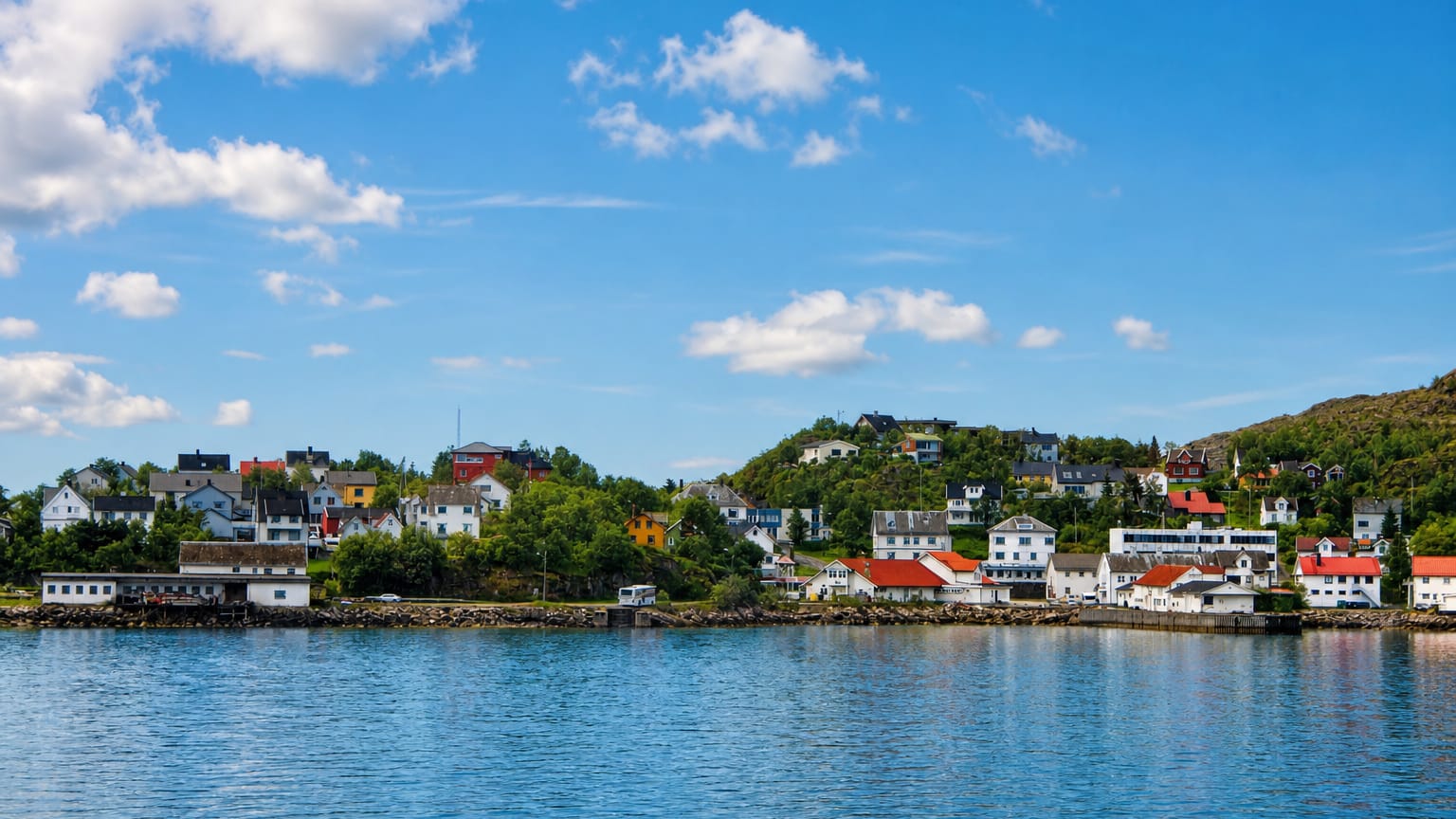 Waterfront of Lødingen, Norway, with white red-roofed houses along the shoreline below a forested hill under blue sky