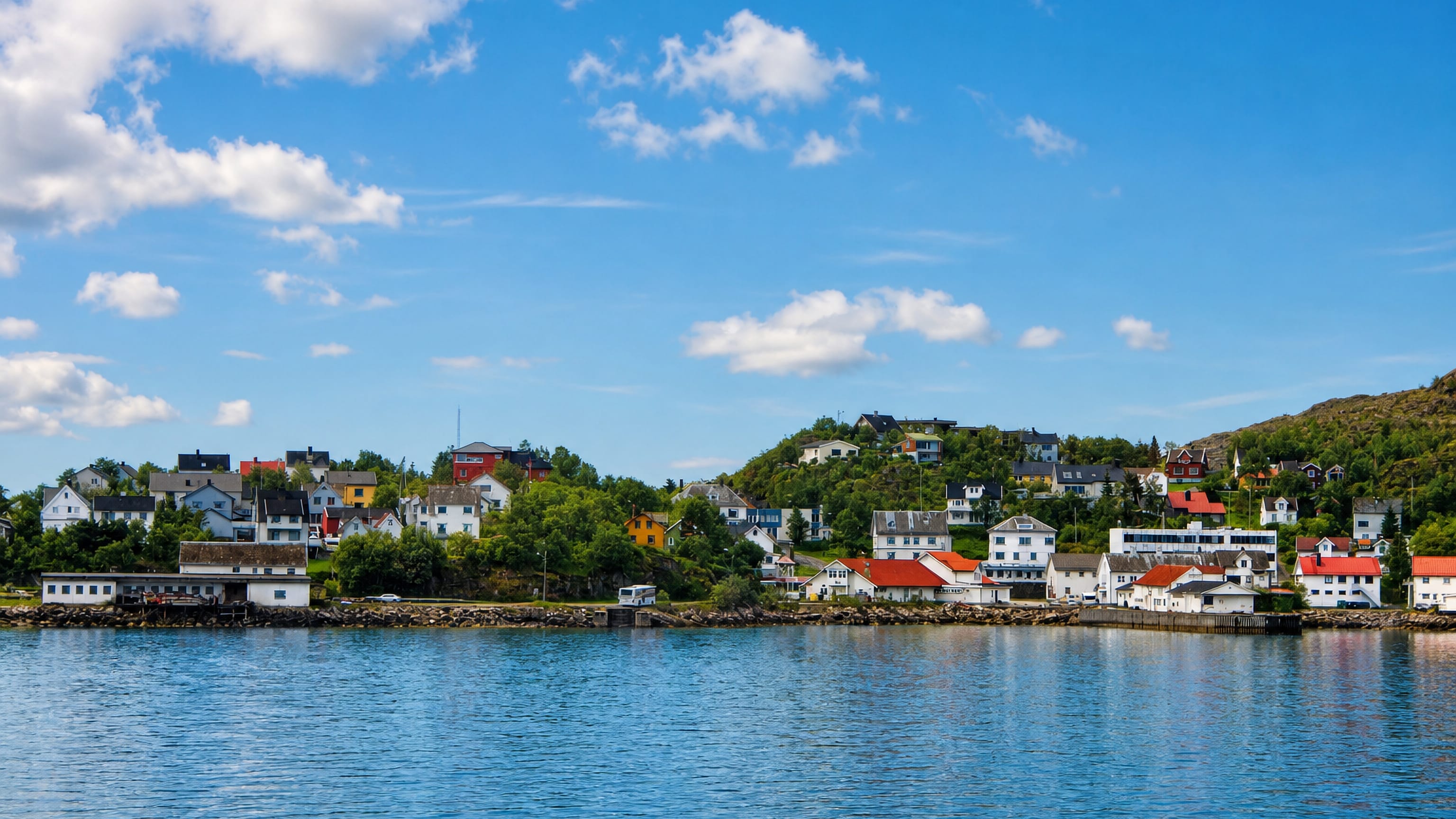 Waterfront of Lødingen, Norway, with white red-roofed houses along the shoreline below a forested hill under blue sky