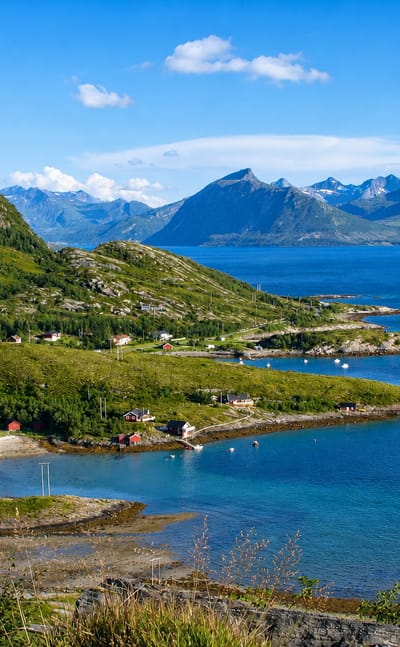Lurøy's green coastal mountains, scattered red and white houses, and a winding road above a blue fjord in Nordland, Norway.