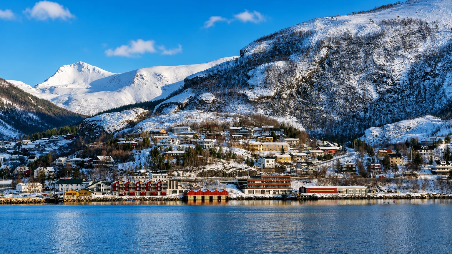 Colourful houses on a snow-covered hillside beneath a fjord peak at the waterfront of Ornes in Nordland, Norway.