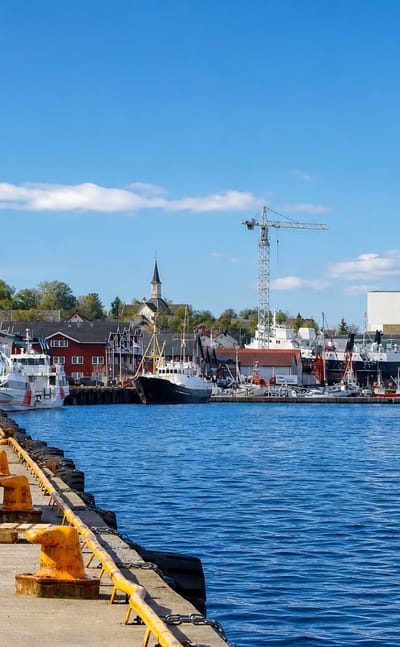 Sandnessjøen harbour in Norway with moored boats, a shipyard crane and red waterfront buildings under a blue sky.