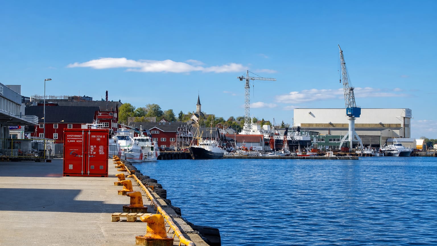 Sandnessjøen harbour in Norway with moored boats, a shipyard crane and red waterfront buildings under a blue sky.