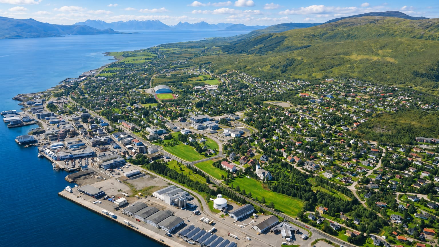 Aerial view of Sortland, Norway, along a coastal sound with harbour quays and green mountains across the water.