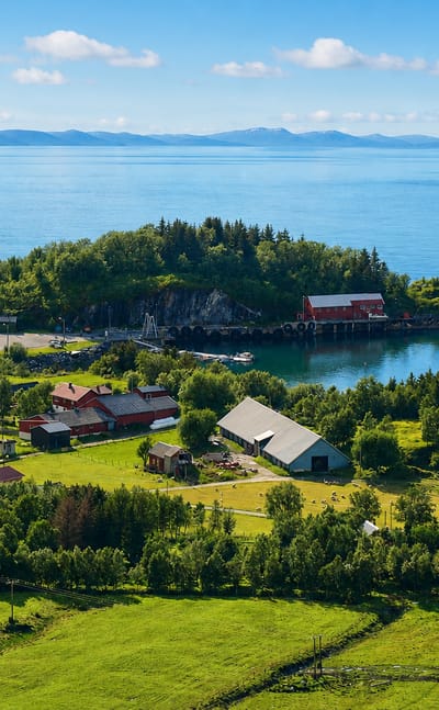 Red farm buildings and green meadows on a wooded headland beside a calm fjord at Vevelstad, Norway.