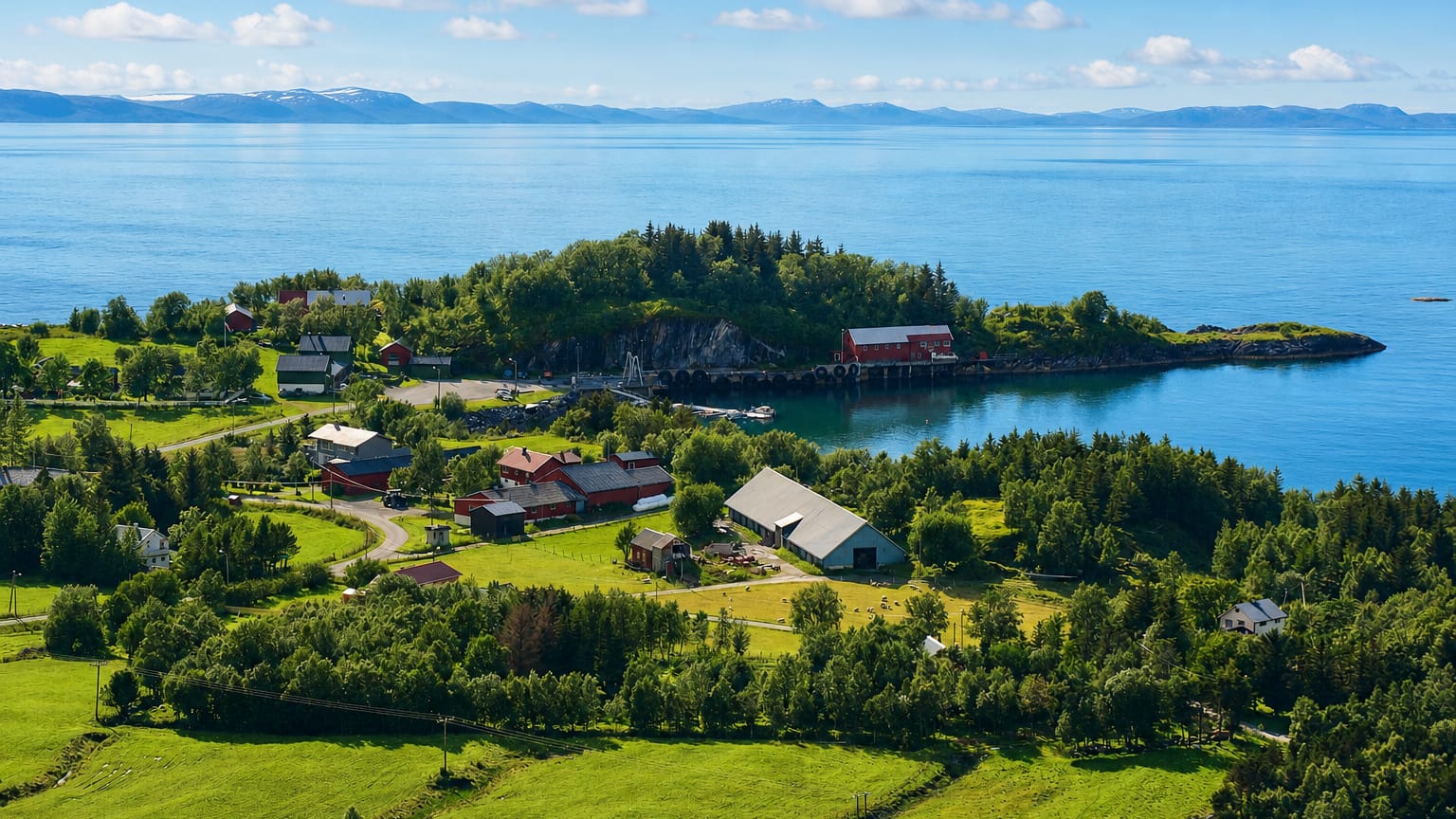 Red farm buildings and green meadows on a wooded headland beside a calm fjord at Vevelstad, Norway.