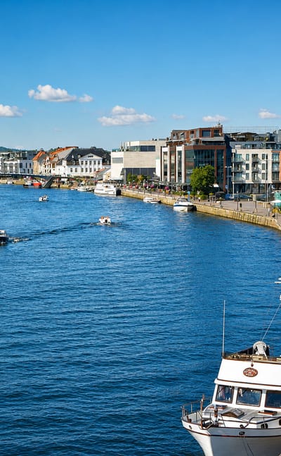 Fredrikstad waterfront on the Glomma river, with moored yachts, a promenade and mixed townhouses under a blue summer sky.