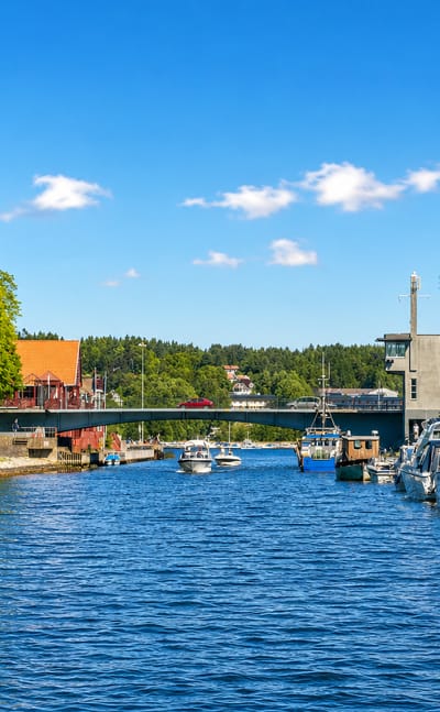 Sailboats moored along the Moss canal by a road bridge, with waterfront townhouses under a bright summer sky.