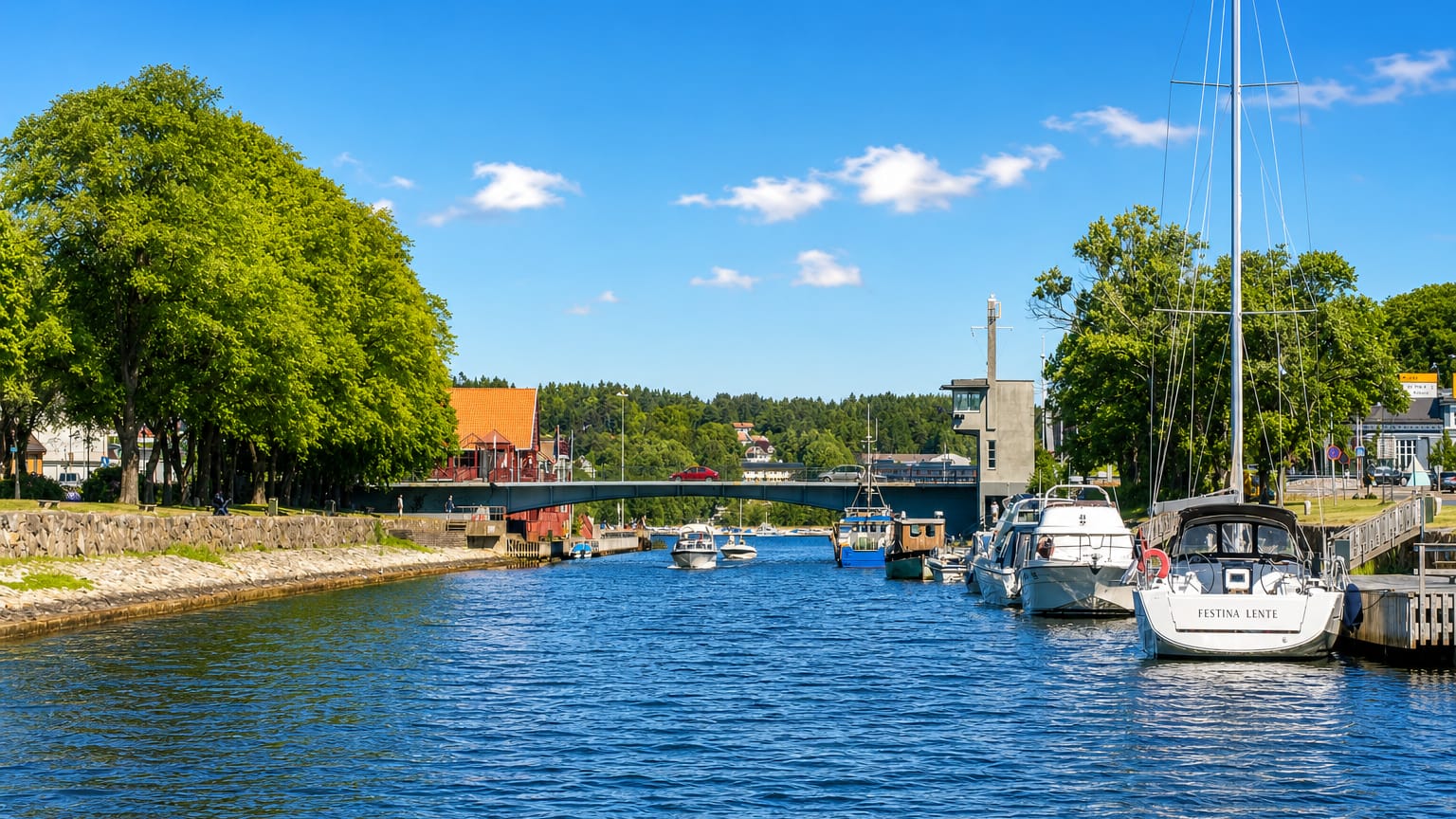 Sailboats moored along the Moss canal by a road bridge, with waterfront townhouses under a bright summer sky.